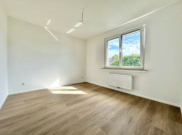 This is a bright, empty room with white walls, light wood flooring, a large window, and a radiator beneath it. Sunlight illuminates one corner.
