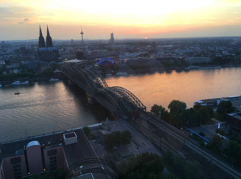 A breathtaking sunset view over Cologne, Germany, featuring the Rhine River, Hohenzollern Bridge, iconic Cathedral, city skyline, and tranquil, softly-lit evening sky.