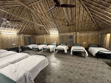 This image shows a rustic bamboo dormitory with multiple single beds, white sheets, and ceiling fans. The room has a cozy, natural, wood-themed ambiance.