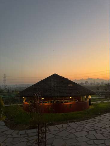 A serene evening scene shows a round, thatched-roof hut illuminated with warm lights, surrounded by greenery, mountains, and a softly glowing sunset sky. Tranquil ambiance prevails.