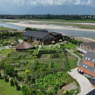 Dieses Bild zeigt einen üppigen grünen Garten, Hütten und ein großes Gebäude an einem Fluss mit entfernten Hügeln, unter einem teilweise bewölkten blauen Himmel. Eine friedliche Szenerie.