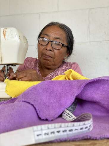 An elderly woman with glasses sews fabric on a sewing machine. She is focused, surrounded by vibrant yellow and purple cloth and a measuring tape.