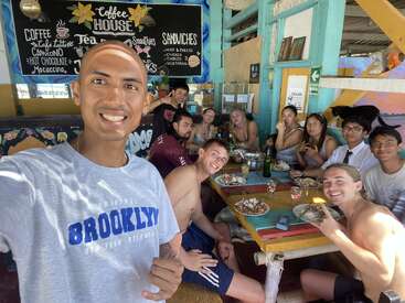 A group of friends enjoys food and drinks together in a colorful, beachside cafe. They smile at the camera, radiating happiness, camaraderie, and vacation vibes.