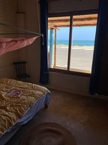 A cozy bedroom with a yellow blanket, mosquito net, and magazine on the bed. Large window reveals a sunny beach, blue sea, and clear sky.