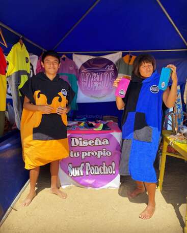 Two people stand barefoot in a colorful market stall, wearing bright surf ponchos. They smile, holding more ponchos. The sign reads: “Diseña tu propio Surf Poncho!”