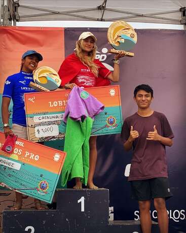 Three winners stand on a podium, holding colorful checks and trophies. The central figure celebrates first place, flanked by smiling competitors against a decorated event backdrop.