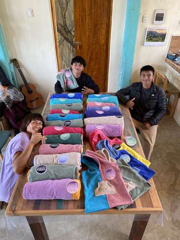 Three smiling people sit around a table displaying colorful, rolled towels with labels. The room is cozy with a guitar, calendar, and wooden accents in the background.