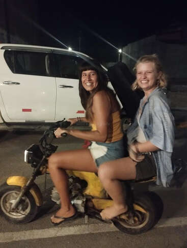 Two young women smile joyfully while sitting on a small yellow motorcycle at night. They appear carefree, dressed casually, enjoying the moment together outdoors.