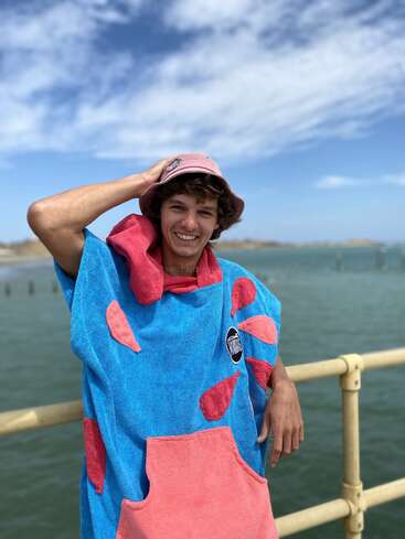 A young man stands smiling on a pier by the water, wearing a colorful blue and pink poncho and bucket hat, under a partly cloudy sky.