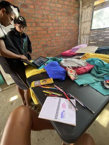Two people stand by a table covered with colorful fabrics. Pencils, a sketchbook with drawings, and a tablet are visible. Bright, rustic workspace atmosphere.