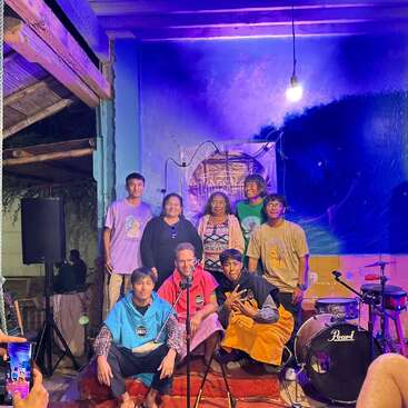 A group of people pose together on a small stage with musical instruments. The background is painted blue, with lights casting a colorful, festive atmosphere.