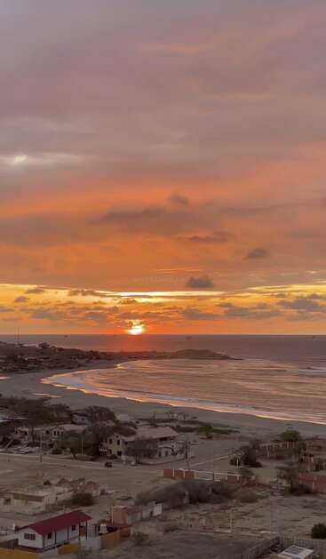 A stunning orange and purple sunset casts a warm glow over a tranquil coastline, reflecting on gentle waves. Quiet houses sit along the peaceful sandy shore.