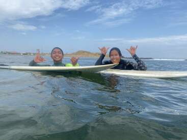 Two people in wetsuits float on surfboards in the ocean, smiling and making shaka hand signs, with a blue sky and distant shoreline behind them.