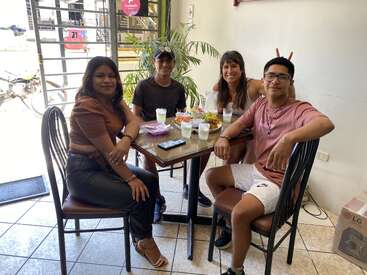 Four friends are sitting around a restaurant table, smiling and enjoying food together. One person playfully makes bunny ears behind another. The atmosphere is casual and friendly.
