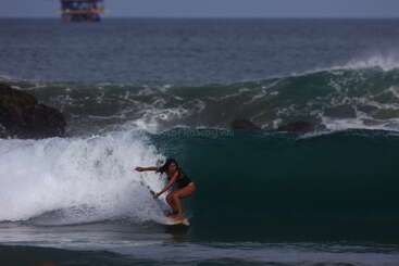 A woman skillfully rides a powerful, dark wave on her surfboard. The ocean appears vast, with an offshore platform visible in the distant background.