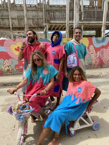 Five people wear colorful poncho towels, posing playfully outdoors. Two ride a tricycle with toys, while others smile. A vibrant mural with hearts and cartoons backgrounds them.