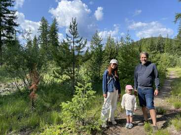 A family of three, smiling, stands on a sunny forest path surrounded by lush green trees under a blue sky with scattered white clouds.