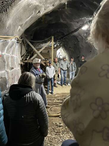 A group of people listens attentively to a guide wearing a helmet inside a dimly lit tunnel, possibly during an educational tour of an underground mine.