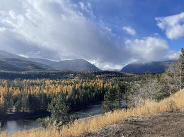 A scenic landscape featuring a river winding through a lush forest of evergreens and golden trees, with majestic mountains and dramatic clouds under a bright blue sky.