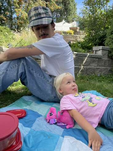 A man in a plaid cap sits on a picnic blanket beside a little girl lying down with a pink stuffed toy dragon, outdoors on a sunny day.