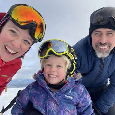 A smiling family of three, wearing winter jackets and ski goggles, poses together outdoors on a snowy day, enjoying time together, likely on a ski trip.