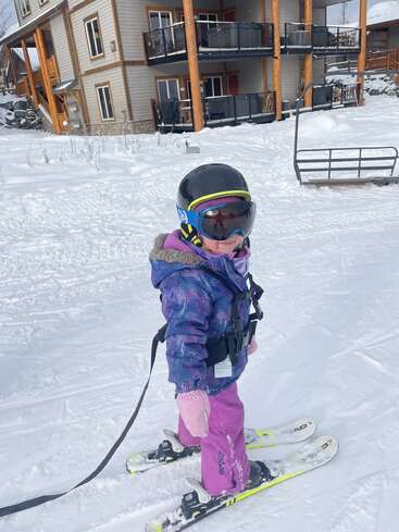 A young child wearing a helmet, goggles, and winter jacket stands on skis in the snow near a lodge, ready for a fun day skiing.