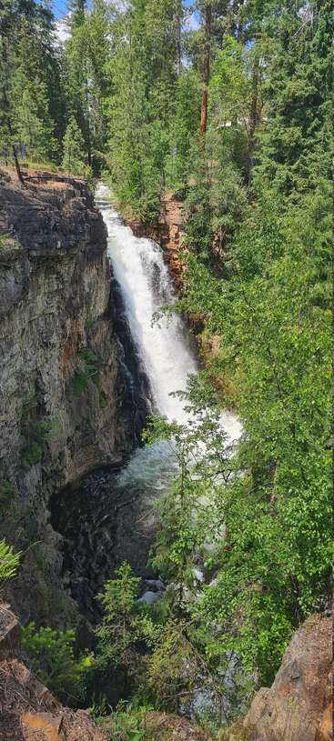 A powerful waterfall cascades down rugged cliffs into a deep pool below, surrounded by lush green forest and tall trees beneath a bright blue sky.