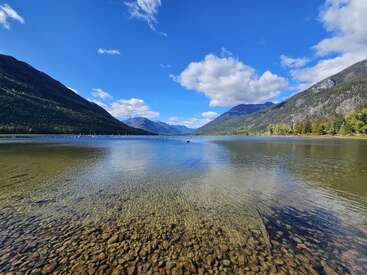 A crystal-clear lake with pebbles beneath its shallow water is nestled between green mountains under a bright blue sky with scattered white clouds. Serene beauty.