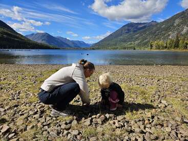 A woman and child crouch on a rocky lakeshore, surrounded by mountains and blue sky, enjoying a peaceful moment in beautiful natural scenery, exploring together.