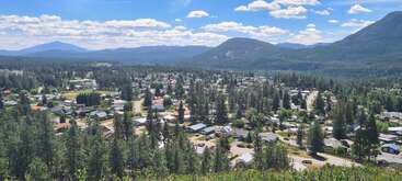 A small town nestled among dense pine forests, surrounded by rolling hills and distant mountains under a bright blue sky with fluffy, scattered white clouds.