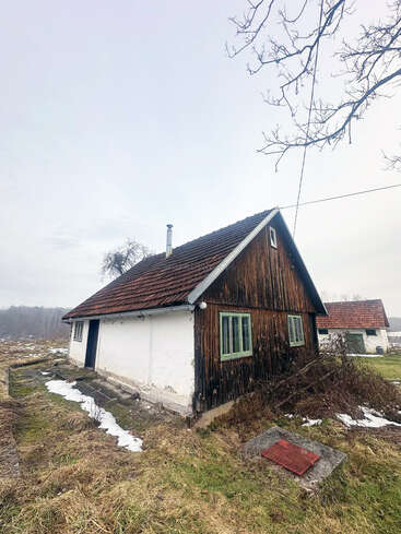 A rustic small house with wooden siding and a tiled roof sits in a grassy, partially snowy field under a gray sky. Bare trees surround the scene.