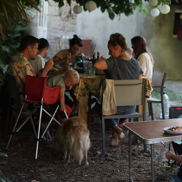 Eine Familie versammelt sich zu einer Mahlzeit im Freien unter einem Laubbaum. Ein Kind interagiert mit einem Hund und schafft eine warme, entspannte Atmosphäre am gemeinsamen Tisch.