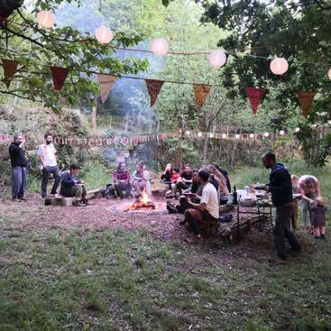 A imagem mostra um grupo de pessoas reunidas em torno de uma fogueira em uma área arborizada, cercadas por árvores e decorações festivas, desfrutando de uma reunião social.