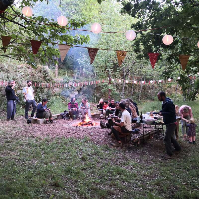 Das Bild zeigt eine Gruppe von Menschen, die um ein Lagerfeuer in einem Waldgebiet versammelt sind, umgeben von Bäumen und festlicher Dekoration, und ein geselliges Beisammensein genießen.