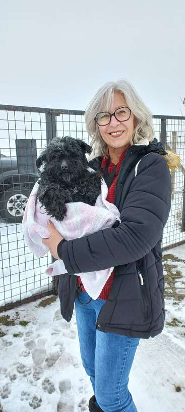 Eine lächelnde Frau mit grauen Haaren und Brille hält an einem verschneiten Tag draußen in der Nähe eines Drahtzauns einen kleinen schwarzen Hund, der in ein pastellfarbenes Handtuch gewickelt ist.