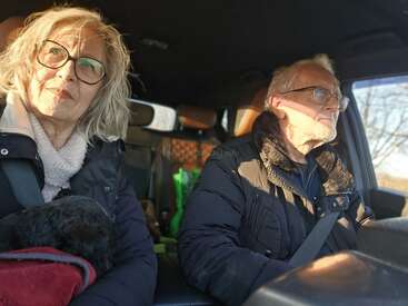 Ein älteres Paar sitzt im Auto, beide mit Brille und Winterjacken, während die Sonne reinscheint. Sie sehen nachdenklich und ruhig aus. Ein Hund ist auch da.