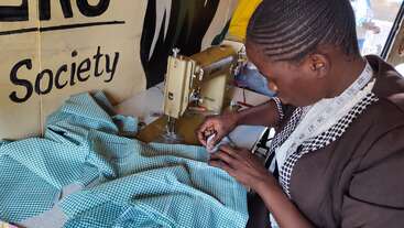 A woman sews green checked fabric using a sewing machine. She is focused on her work and wears a measuring tape around her neck in a workshop setting.