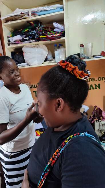 Two women are happily interacting in a room filled with colorful fabrics and shelves. One is smiling while applying makeup to the other, creating a joyful moment.