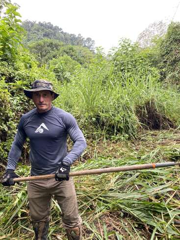 Un homme portant un équipement de plein air et des gants se tient dans une jungle verte et dense, tenant un long poteau en bois, entouré d'herbes hautes et de feuillage épais.