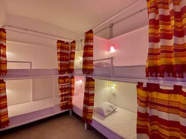 L'image représente une pièce compacte avec quatre lits superposés, chacun avec des draps blancs et des rideaux à rayures rouges et jaunes, éclairés par de petites lumières sur les murs.