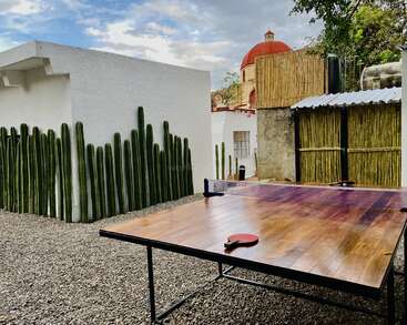 L'image représente un cadre extérieur serein avec une table de ping-pong située dans une zone de gravier, entourée d'un bâtiment blanc et d'une clôture ornée de cactus.