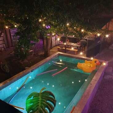 La imagen representa una serena escena nocturna en el patio trasero de la piscina, con un patito de goma amarillo flotando, rodeado de luces y frondosos árboles, creando un ambiente tranquilo.