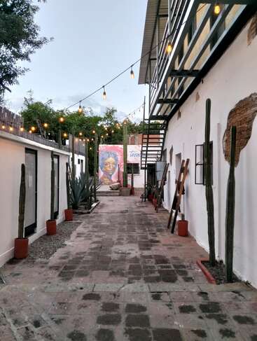 Une ruelle rustique avec des guirlandes lumineuses, des cactus en pot le long des murs, des bâtiments blancs et une fresque vibrante représentant le visage d'une femme à la fin. Atmosphère paisible.