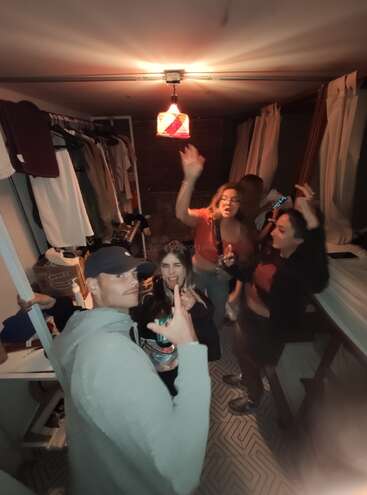 Cinq amis s'amusent dans une petite pièce confortable. Ils dansent, posent et sourient sous une lumière chaude, entourés de vêtements et de lits superposés.