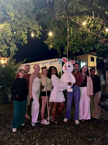 Un groupe de huit personnes pose ensemble la nuit, sous des guirlandes lumineuses, en plein air, près d'arbres. Elles sourient et présentent une grande piñata animale colorée.