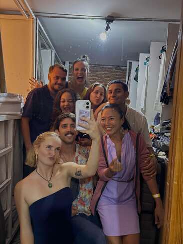 Un groupe de huit amis pose étroitement ensemble, souriant et faisant des grimaces pour un selfie dans un miroir, dans une pièce confortable et bien éclairée au décor décontracté.