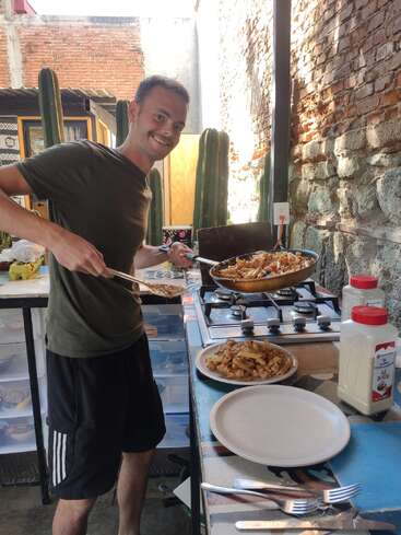 Un homme souriant se tient dans une cuisine en plein air, servant des pâtes d'une casserole à une assiette. Des cactus et des murs de briques rustiques forment une toile de fond accueillante et ensoleillée.