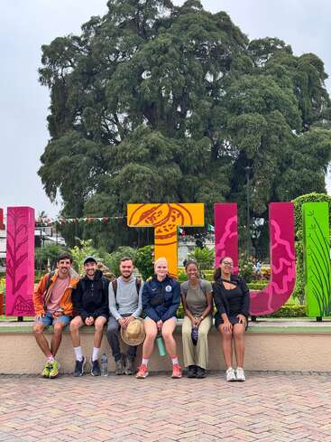Six personnes sont assises et sourient sur un rebord devant des lettres colorées "TULE", avec de la verdure luxuriante et un grand arbre bien en évidence à l'arrière-plan.