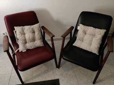 Deux fauteuils vintage, l'un rouge et l'autre noir, sont placés côte à côte. Chacun est orné d'un coussin décoratif beige à motifs floraux, devant un mur uni.