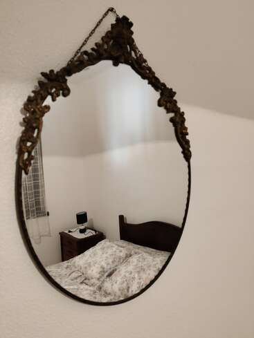 Un miroir ovale richement décoré reflète une chambre à coucher chaleureuse, avec un lit à motifs floraux, une tête de lit en bois, une table de chevet, une lampe et une fenêtre ornée d'un rideau clair. Une ambiance chaleureuse.